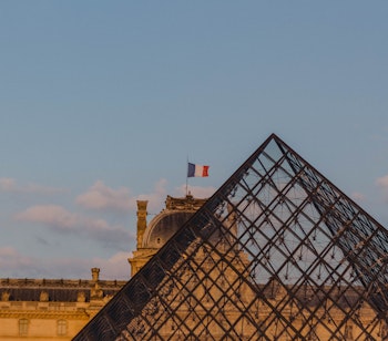 Den ikoniske glaspyramide ved Louvre-museet i Paris med det historiske palads og det franske flag mod en blå himmel
