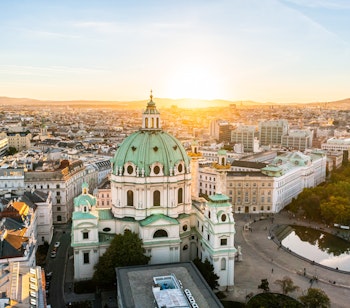 Luftfoto af St. Charles Kirke i Wien ved solnedgang, der fremhæver barokarkitektur og bybillede.