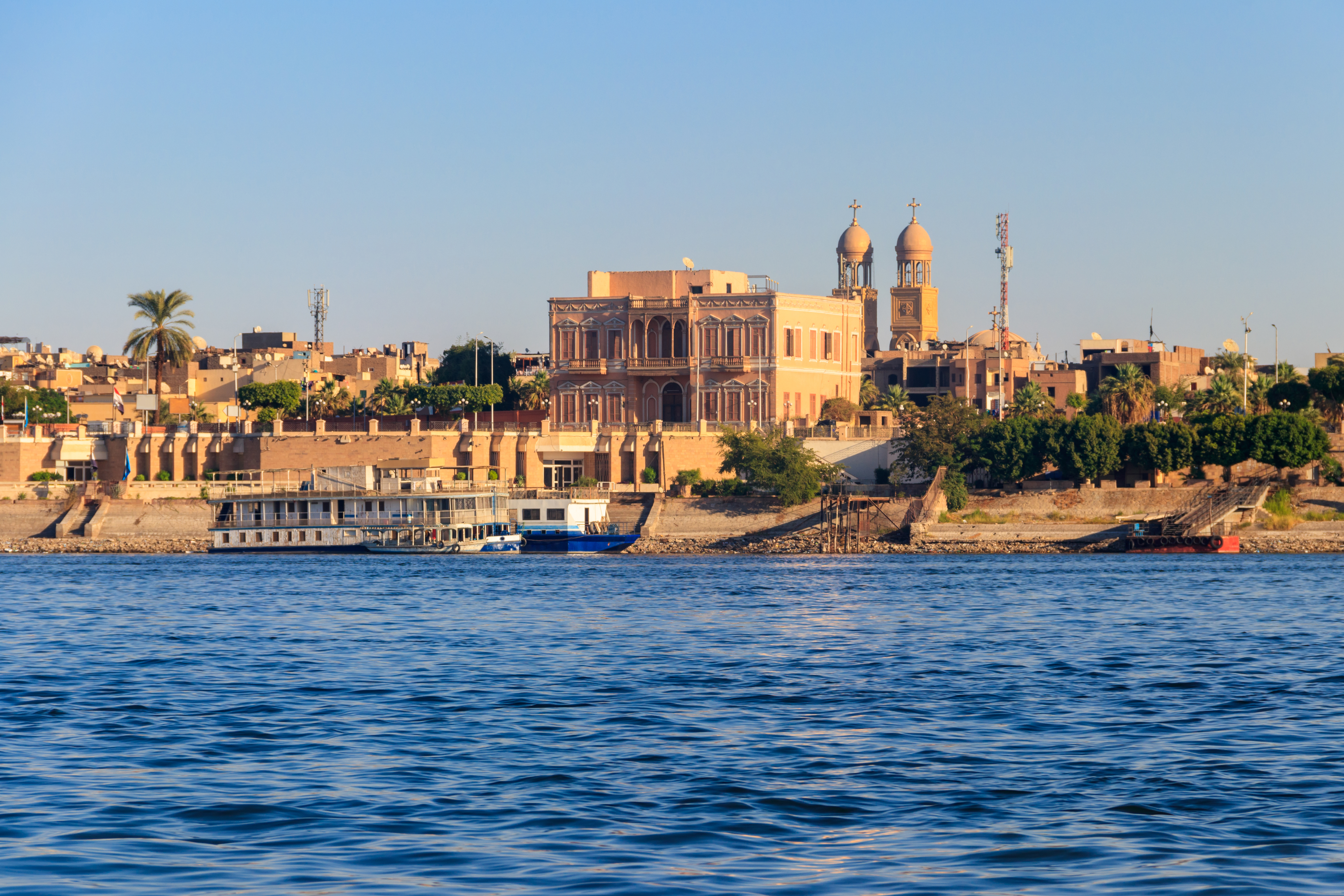 Udsigt over Luxors skyline langs Nilen med traditionel egyptisk arkitektur, moskékupler og et krydstogtskib ved bredden i gyldent aftenlys