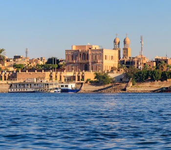 Udsigt over Luxors skyline langs Nilen med traditionel egyptisk arkitektur, moskékupler og et krydstogtskib ved bredden i gyldent aftenlys