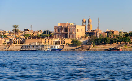 Udsigt over Luxors skyline langs Nilen med traditionel egyptisk arkitektur, moskékupler og et krydstogtskib ved bredden i gyldent aftenlys