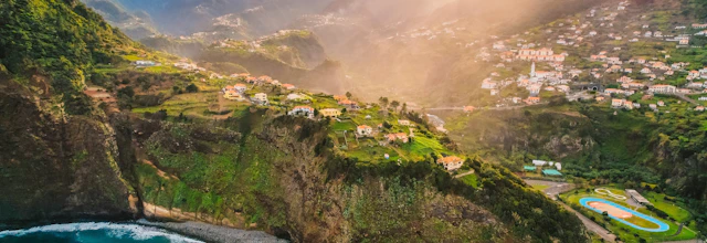 Betagende luftfoto fra Guindaste-udsigtspunktet på Madeira med dramatiske klipper, azurblåt hav og idylliske bjerglandsbyer badet i gyldent sollys