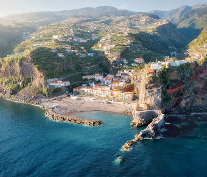 Betagende udsigt over den maleriske kystby Ponta do Sol på Madeira under solnedgang, med farverige huse på klipperne og det blå Atlanterhav badet i gyldent aftenlys