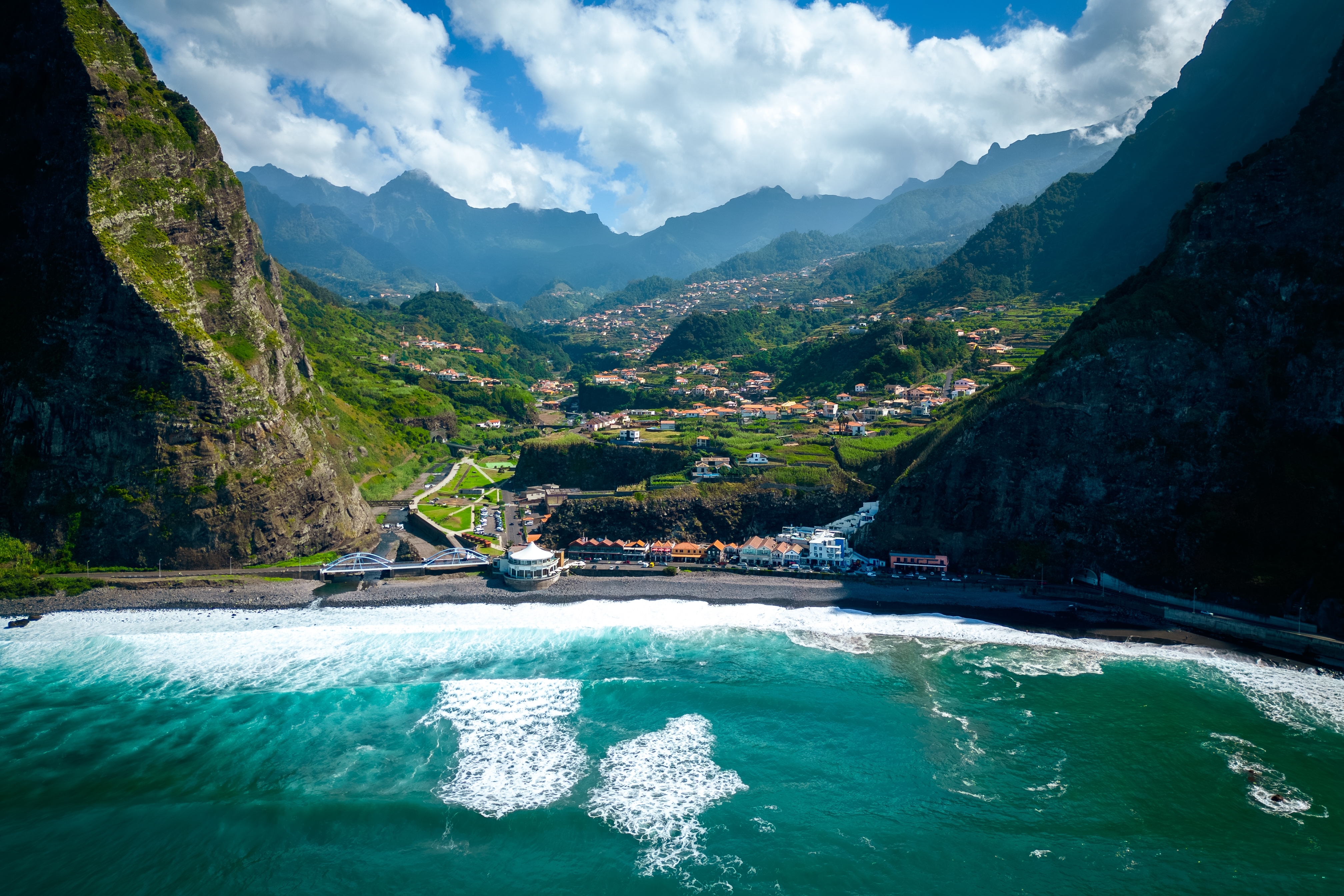 Betagende luftfoto af São Vicente på Madeira med sort sandstrand, brusende atlanterhavsbølger og dramatiske bjerge i baggrunden