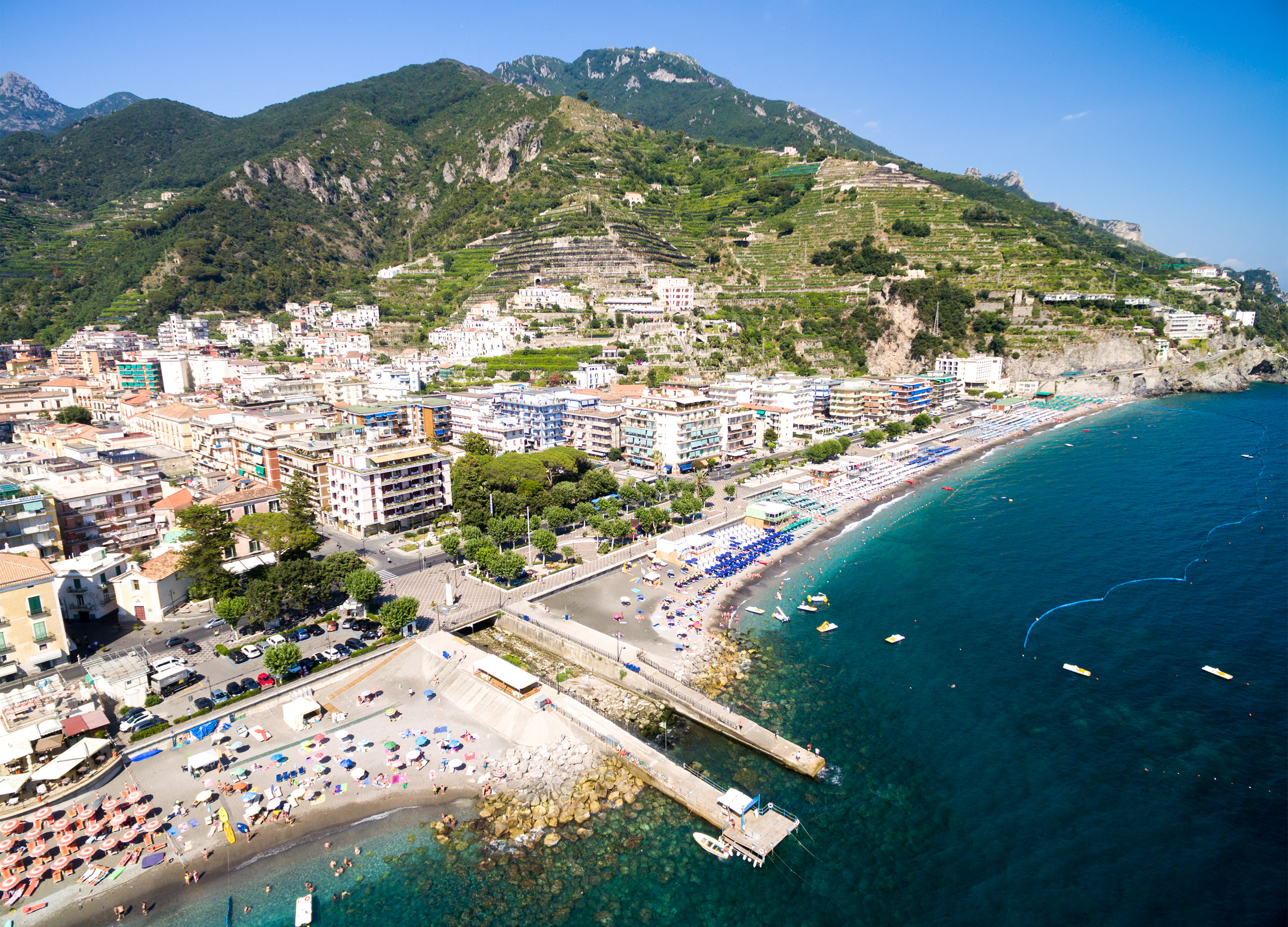 Luftfoto over Maiori på Amalfikysten i Italien med turkisblåt Middelhav, sandstrand med farverige parasolder og grønne terrассerede bjergsider