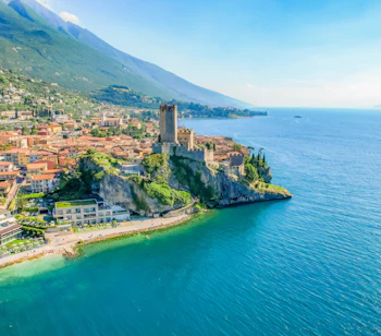 Luftfoto af Scaliger-slottet på klippen ved den turkisblå Gardasø i Malcesine, Italien, med alperne i baggrunden