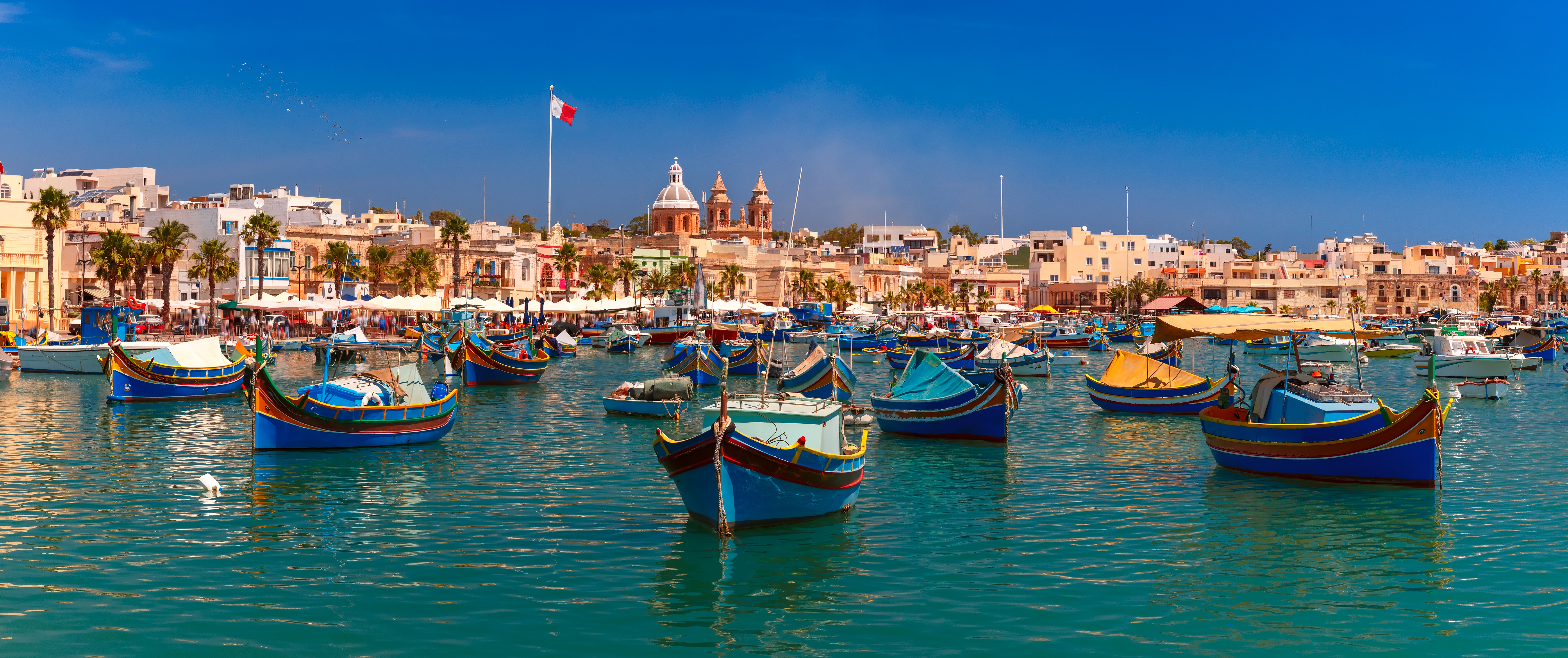 Farverige traditionelle fiskerbåde i Marsaxlokk havn på Malta med historiske bygninger og kirketårne i baggrunden
