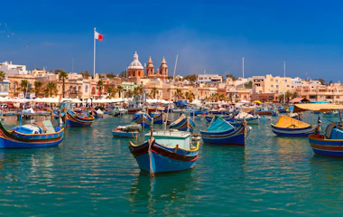 Farverige traditionelle fiskerbåde i Marsaxlokk havn på Malta med historiske bygninger og kirketårne i baggrunden