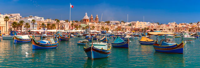 Farverige traditionelle fiskerbåde i Marsaxlokk havn på Malta med historiske bygninger og kirketårne i baggrunden