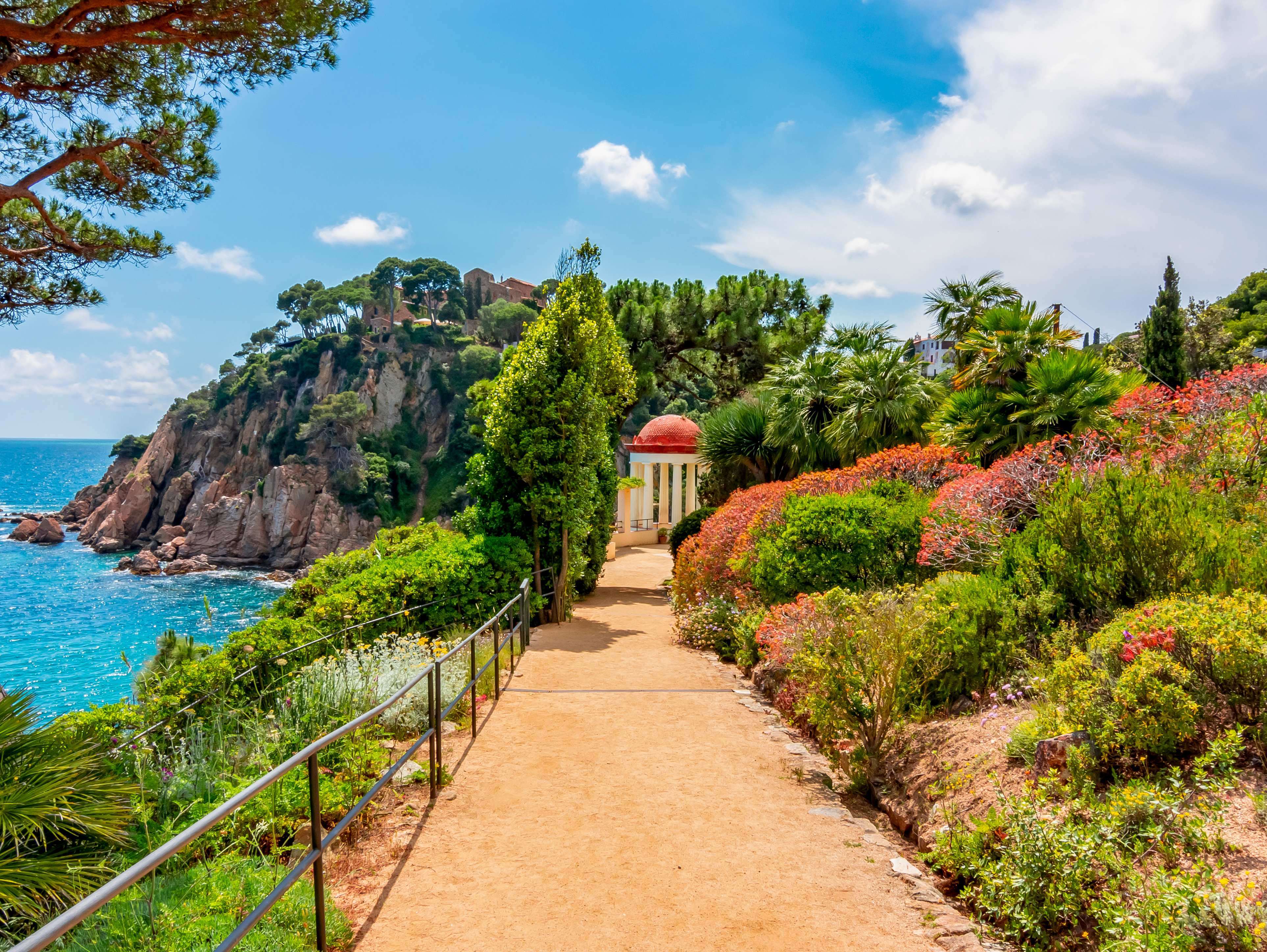Smuk havesti i Marimurtra Botanisk Have i Blanes på Costa Brava med hvid pavillon, frodige planter og turkisblåt middelhav i baggrunden