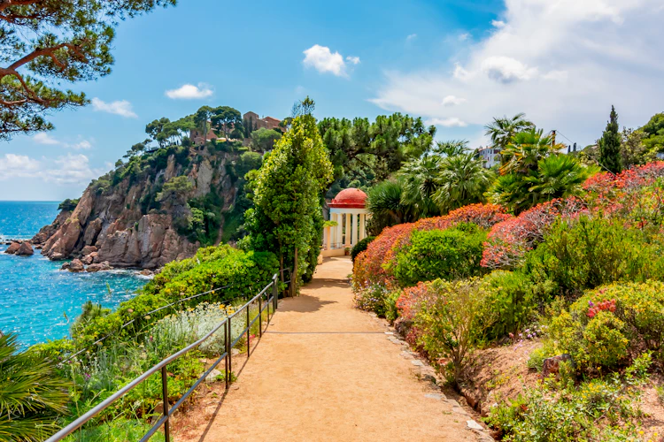 Smuk havesti i Marimurtra Botanisk Have i Blanes på Costa Brava med hvid pavillon, frodige planter og turkisblåt middelhav i baggrunden