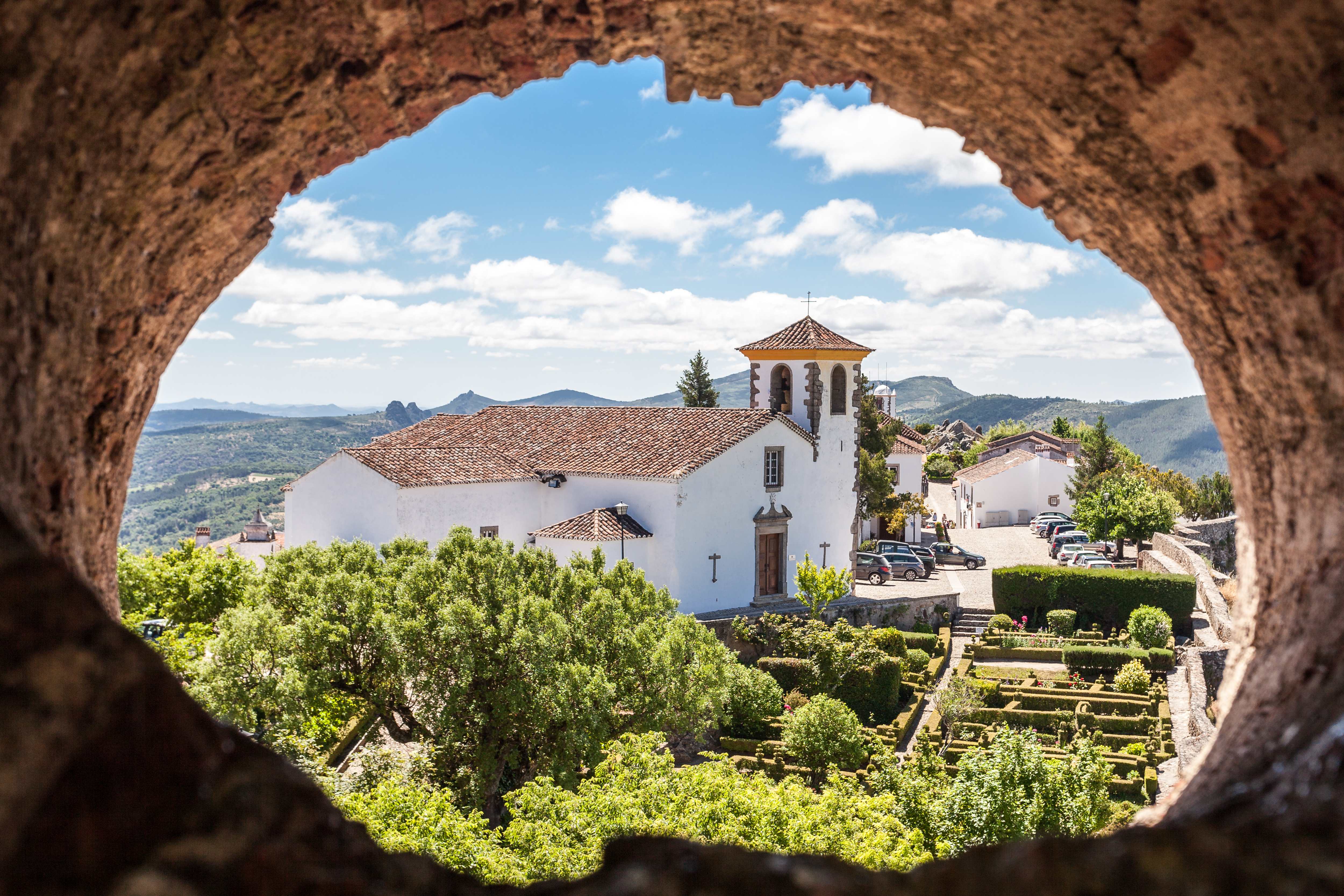 Udsigt gennem hul i stenmur til den historiske hvide kirke Santa Maria i Marvão, Portugal, omgivet af grønne haver og bjerglandskab