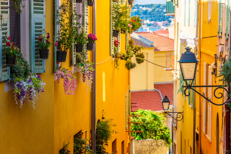 Smuk smal gade i Mentons gamle bydel med gule facader, hængende blomsterkurve og vintage gadelygter på den franske Riviera