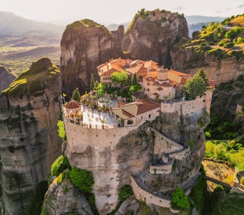 Luftfoto af Meteora-klostrene bygget på høje klippeformationer i Grækenland, der viser Det Hellige Meteoron-kloster og Varlaam med terrakottatage mod grønne bjerge