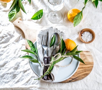 Elegant middelhavsinspireret bordopdækning med hvid tallerken, bestik og appelsingren på hvid linneddug