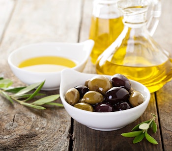 Frisk olivenolie i glasflaske med sorte og grønne oliven og olivenblade arrangeret på et rustikt træbord, typisk for middelhavskøkkenet