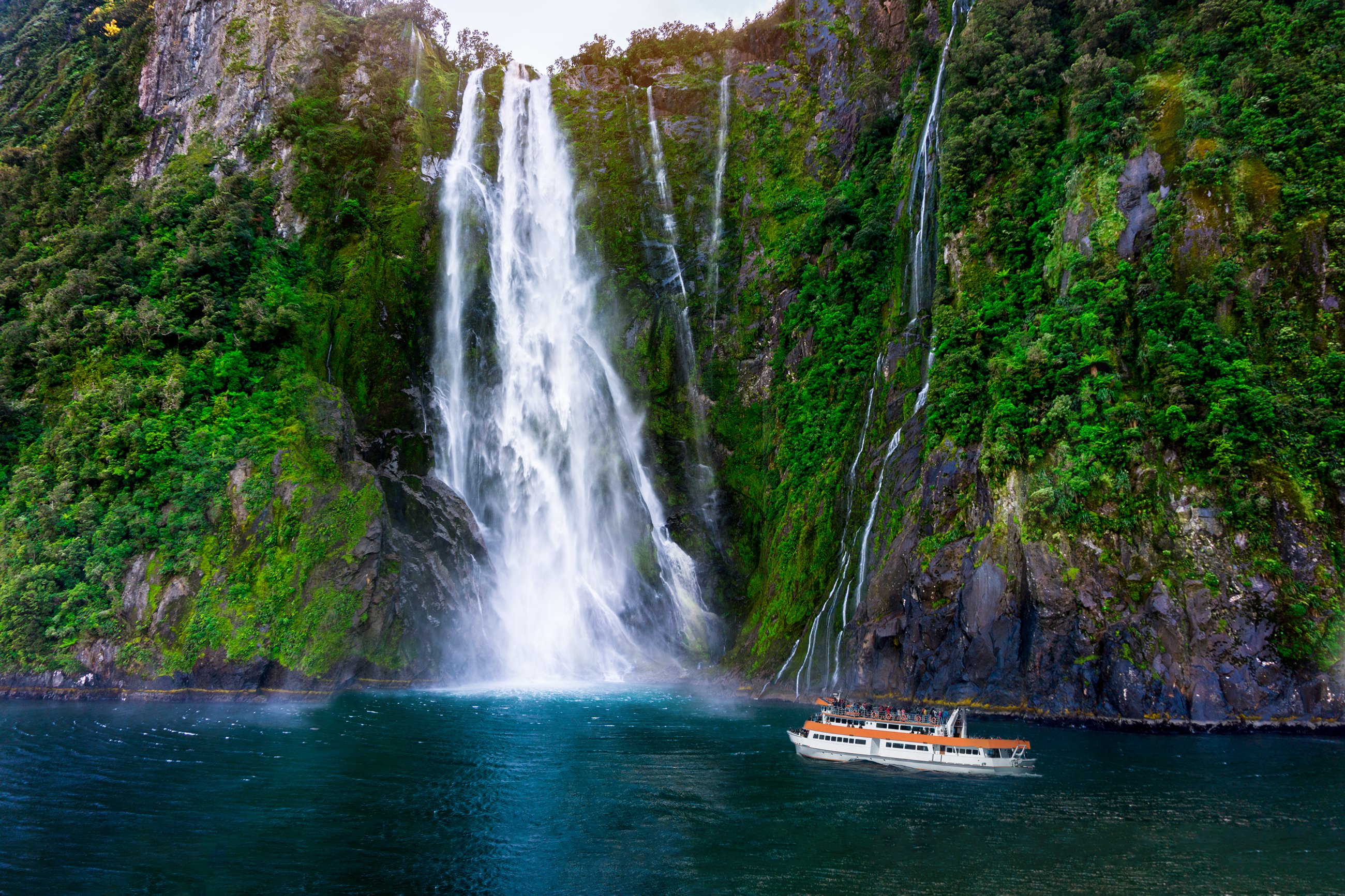 Turistfaerge naermer sig det majestætiske Stirling Falls, der styrter ned ad stejle grønne klipper i Milford Sound, omgivet af dramatisk bjerglandskab i Fiordland National Park, New Zealand