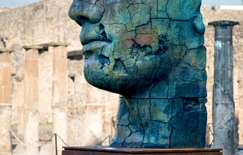 Ikonisk blå bronzeskulptur af Igor Mitoraj forestillende et ansigt ved de antikke ruiner i Pompeji, Italien, med oldtidsarkitektur i baggrunden