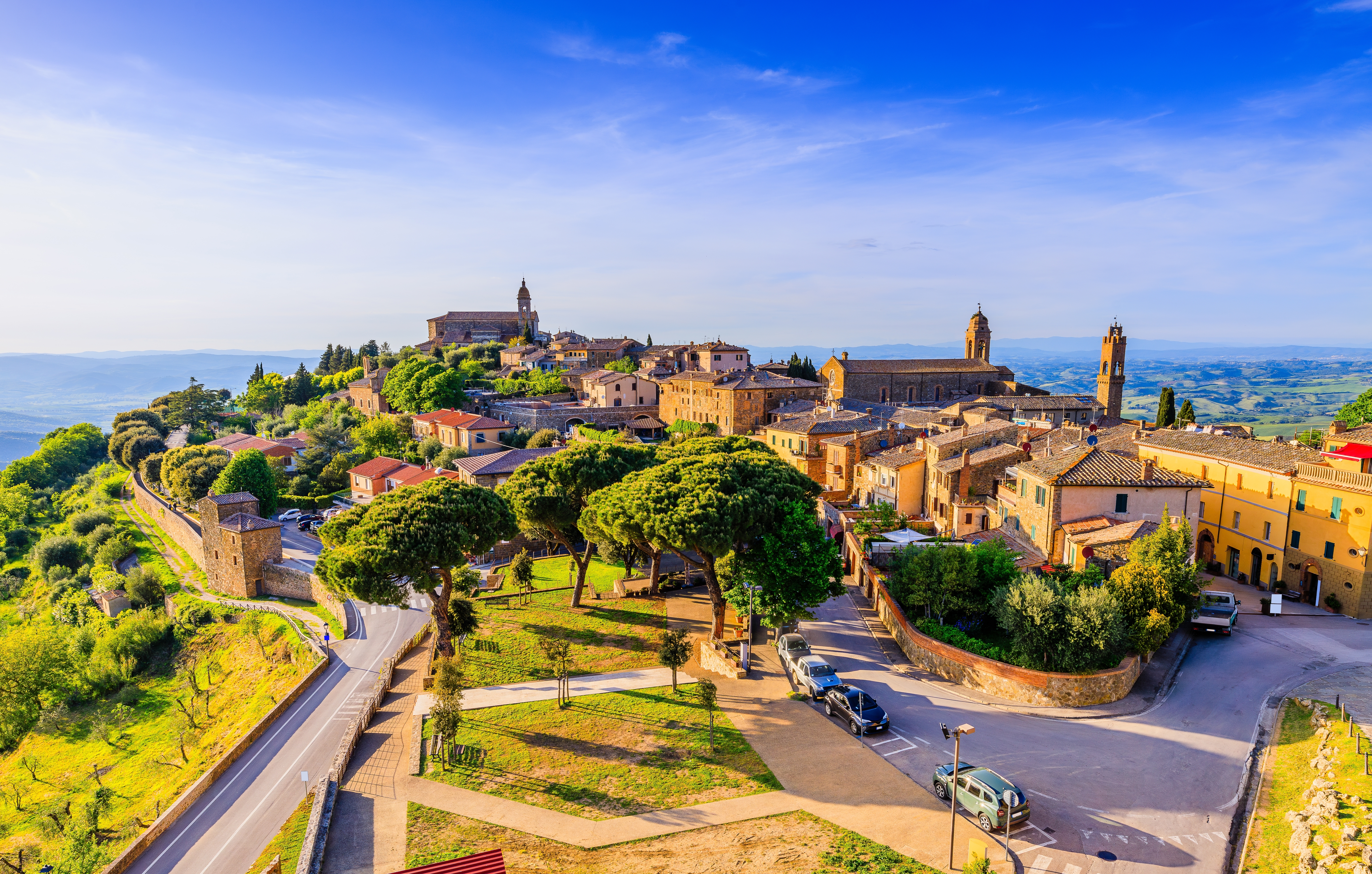 Luftfoto af den middelalderlige by Montalcino i Toscana, Italien, med historiske stenbygninger, kirketårne og det grønne toscanske landskab under en blå himmel