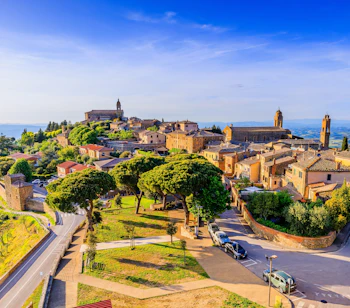 Luftfoto af den middelalderlige by Montalcino i Toscana, Italien, med historiske stenbygninger, kirketårne og det grønne toscanske landskab under en blå himmel