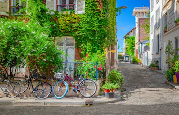 Charmerende brolagt gade i Montmartre, Paris, med vedbend-dækkede bygninger og parkerede cykler langs et blåt jerngelænder på en solrig sommerdag