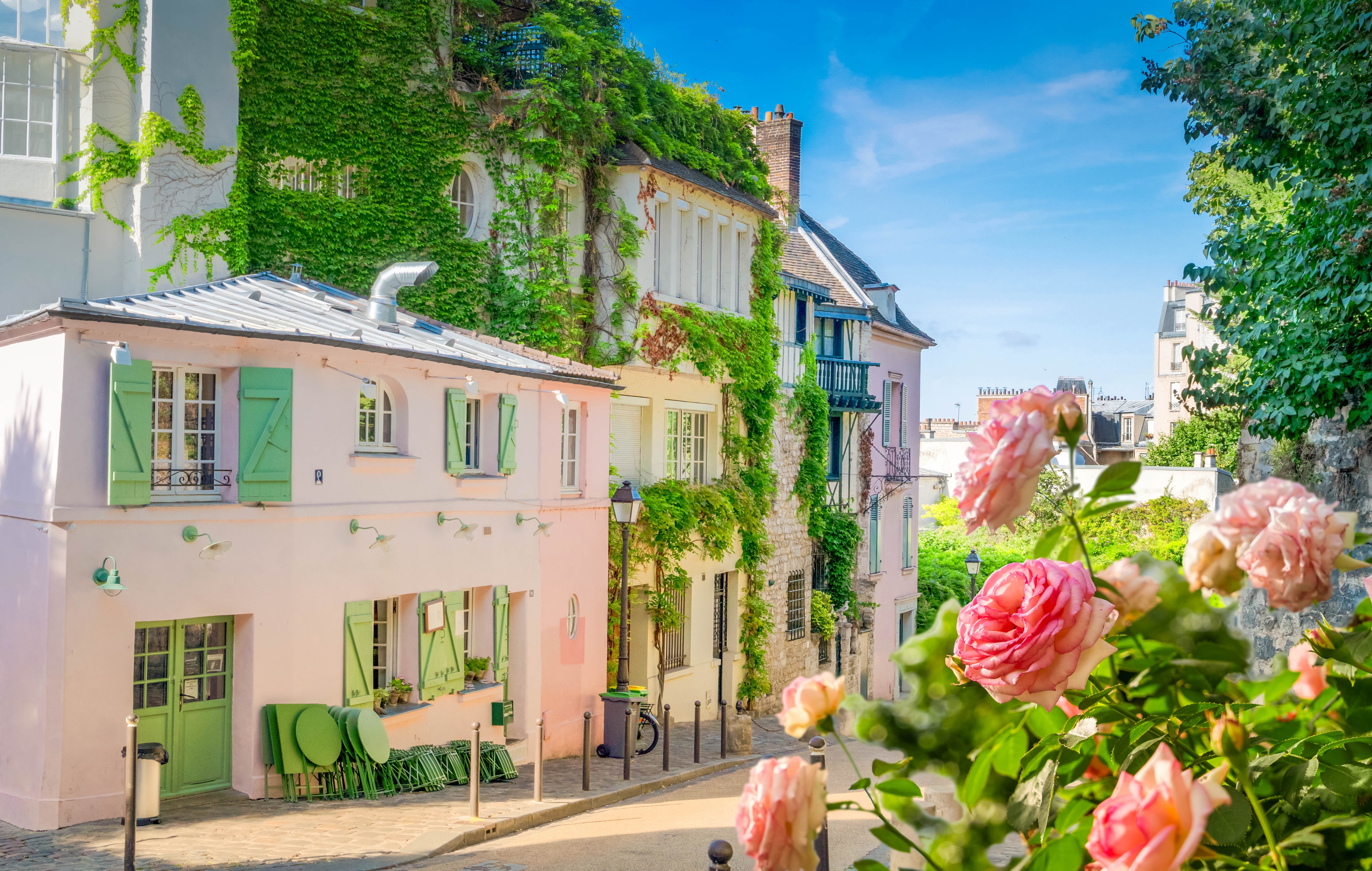 Hyggelig gade i Montmartre, Paris, med lyserøde bygninger, grønne skodder og blomstrende roser i forgrunden på en smuk sommerdag