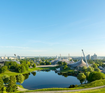 Betagende morgenudsigt over Olympiaparken i München med det ikoniske Olympiatårn, frodig grøn natur og den smukke sø omgivet af Bayerns hovedstads skyline