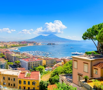 Betagende udsigt over Napoli-bugten fra Posillipo-højderne med Vesuv-vulkanen i baggrunden og karakteristiske pinjetræer i forgrunden på en solrig dag