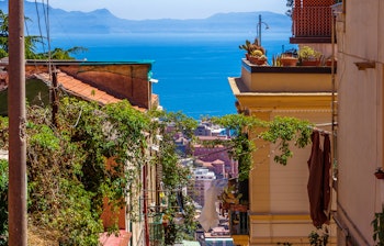 Charmerende smal gade i det historiske Quartieri Spagnoli-kvarter i Napoli med farverige bygninger, balkoner med kaktusser og udsigt til den blå Napoli-bugt