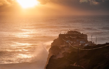 Betagende solnedgang ved fyrtårnet i Nazaré, Portugal, med imponerende bølger fra Atlanterhavet der slår mod kysten