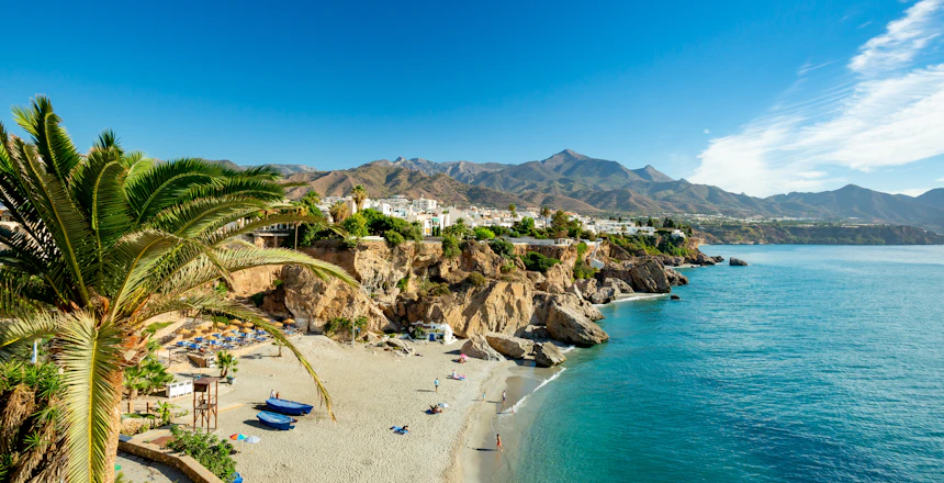 Idyllisk sandstrand ved Nerja i Andalusien med turkisblåt hav, hvide bygninger på klipperne og bjerge i baggrunden