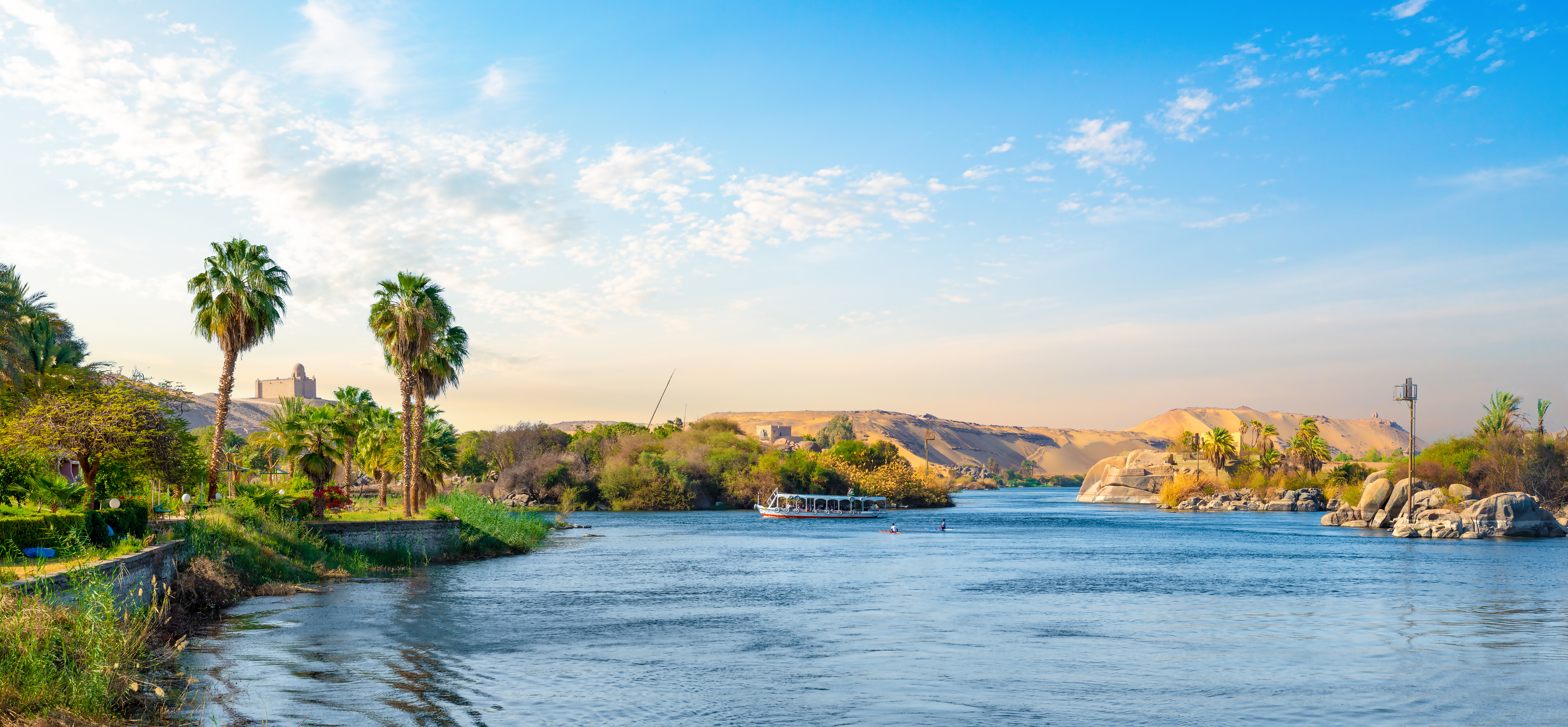 Panoramaudsigt over Nilen i Aswan, Egypten, med palmetræer langs bredden, ørkenklitter og en båd på det rolige blå vand under en klar himmel