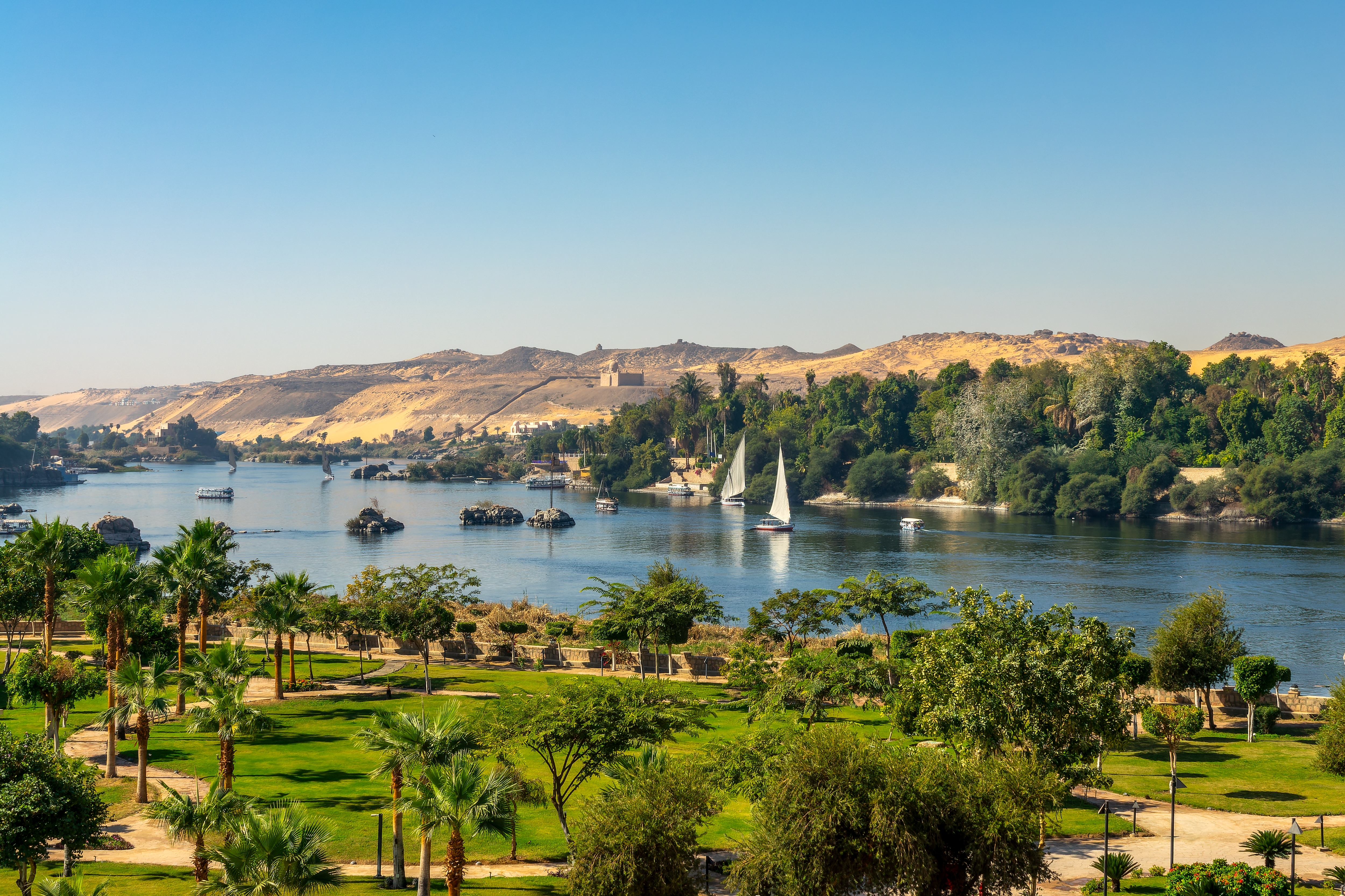 Betagende luftpanorama over Nilen ved Aswan i Egypten, hvor traditionelle felucca-sejlbåde med hvide sejl navigerer de blå vande omgivet af klippeformationer og ørkenlandskab