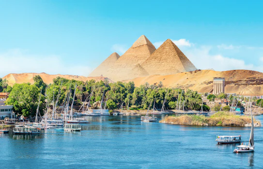 Panoramaudsigt over Nilen i Aswan med traditionelle felucca-sejlbåde, frodige palmtræer og de ikoniske egyptiske pyramider i baggrunden under en klar blå himmel