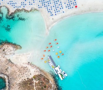 Luftfoto af Nissi Beach i Ayia Napa, Cypern med turkisblåt vand, hvid sandstrand og blå strandparasoller