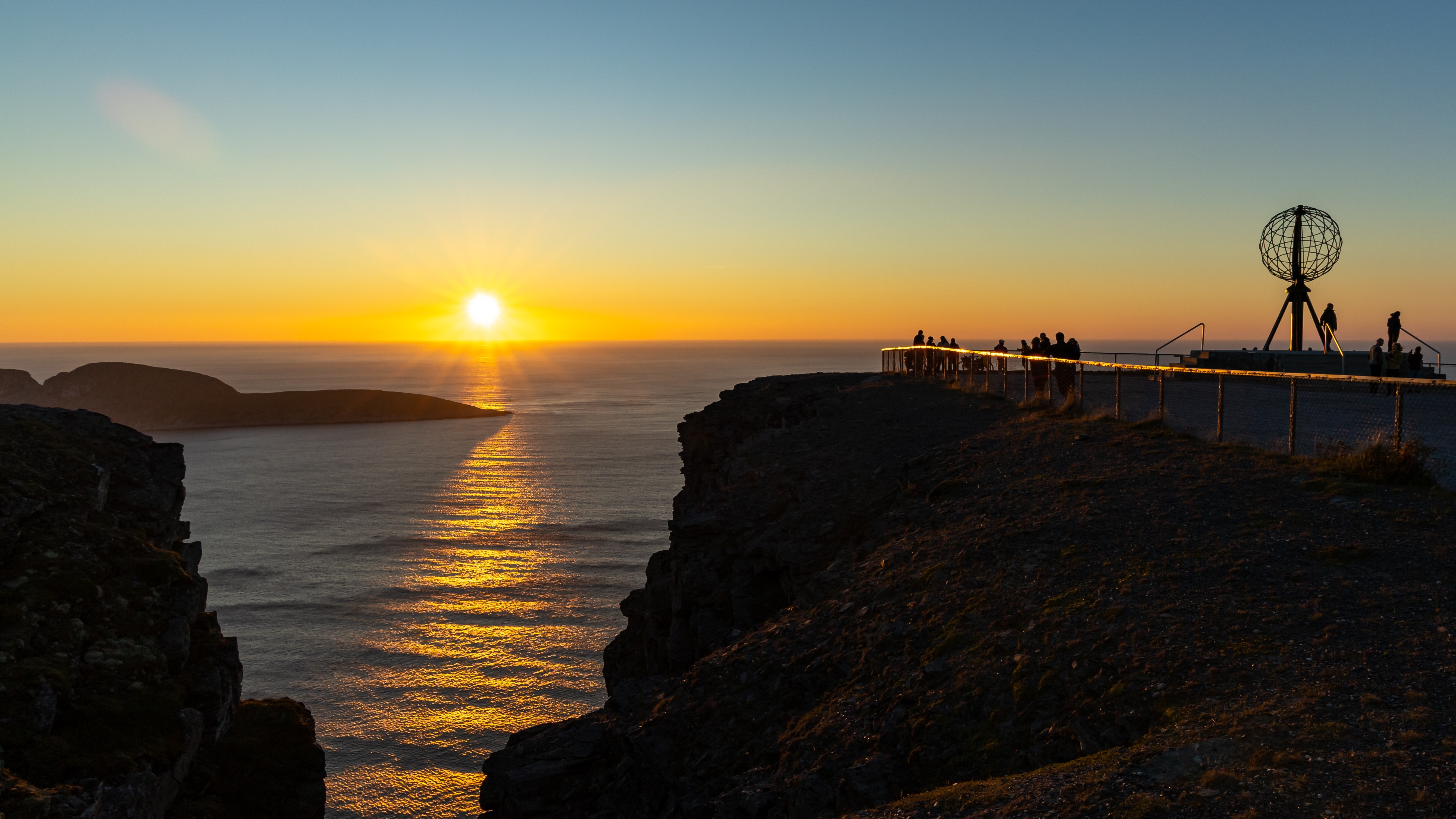 Magisk solnedgang ved Nordkapp i Norge med det ikoniske globus-monument på klippen og den gyldne sol, der spejler sig i Ishavet