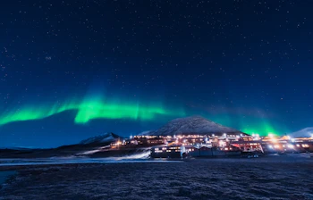 Nordlys og aurora borealis lyser grønt op over den oplyste by Longyearbyen på Svalbard i Norge med snedækkede bjerge og stjerneklar nattehimmel