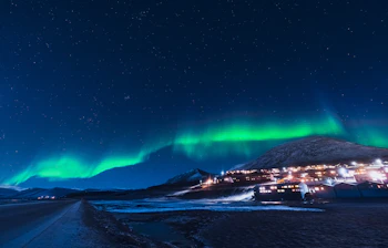 Grønne nordlys over arktisk landsby med sneklædte bjerge under stjerneklar nat