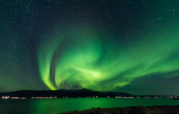 Spektakulært nordlys i grønne nuancer danser over arktisk landskab med stjerneklar himmel og vandspejling