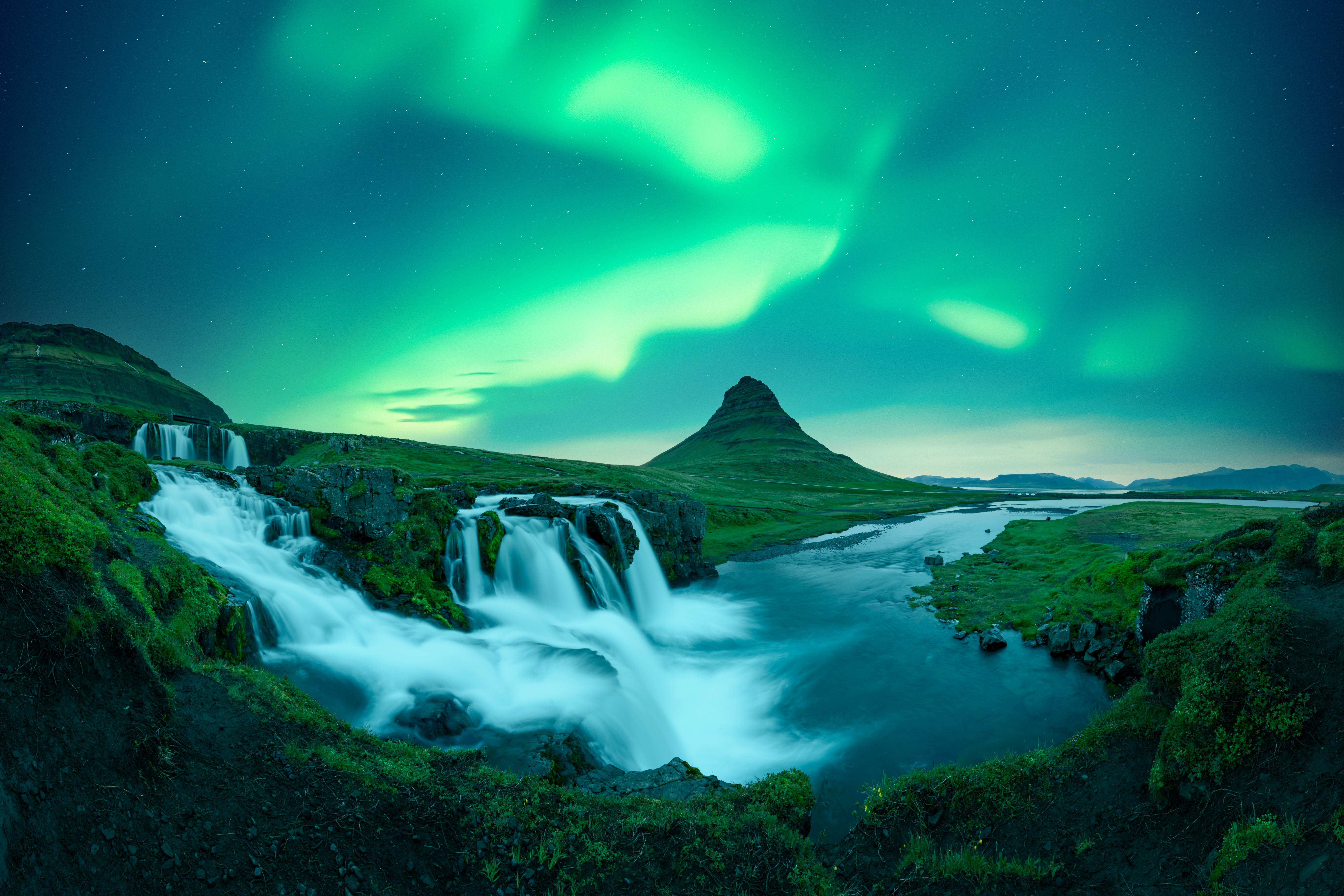 Magiske nordlys danser over Kirkjufellsfoss-vandfaldet med det ikoniske kegleformede Kirkjufell-bjerg i baggrunden, et af Islands mest fotograferede naturfænomener