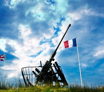 Historisk kanon ved Gold Beach i Arromanches, Normandiet med franske og britiske flag under dramatisk himmel - mindested for D-dag landingen