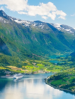 Imponerende krydstogtskib der sejler gennem en storslået norsk fjord med høje bjerge og perfekte spejlinger i det rolige, turkisblå vand