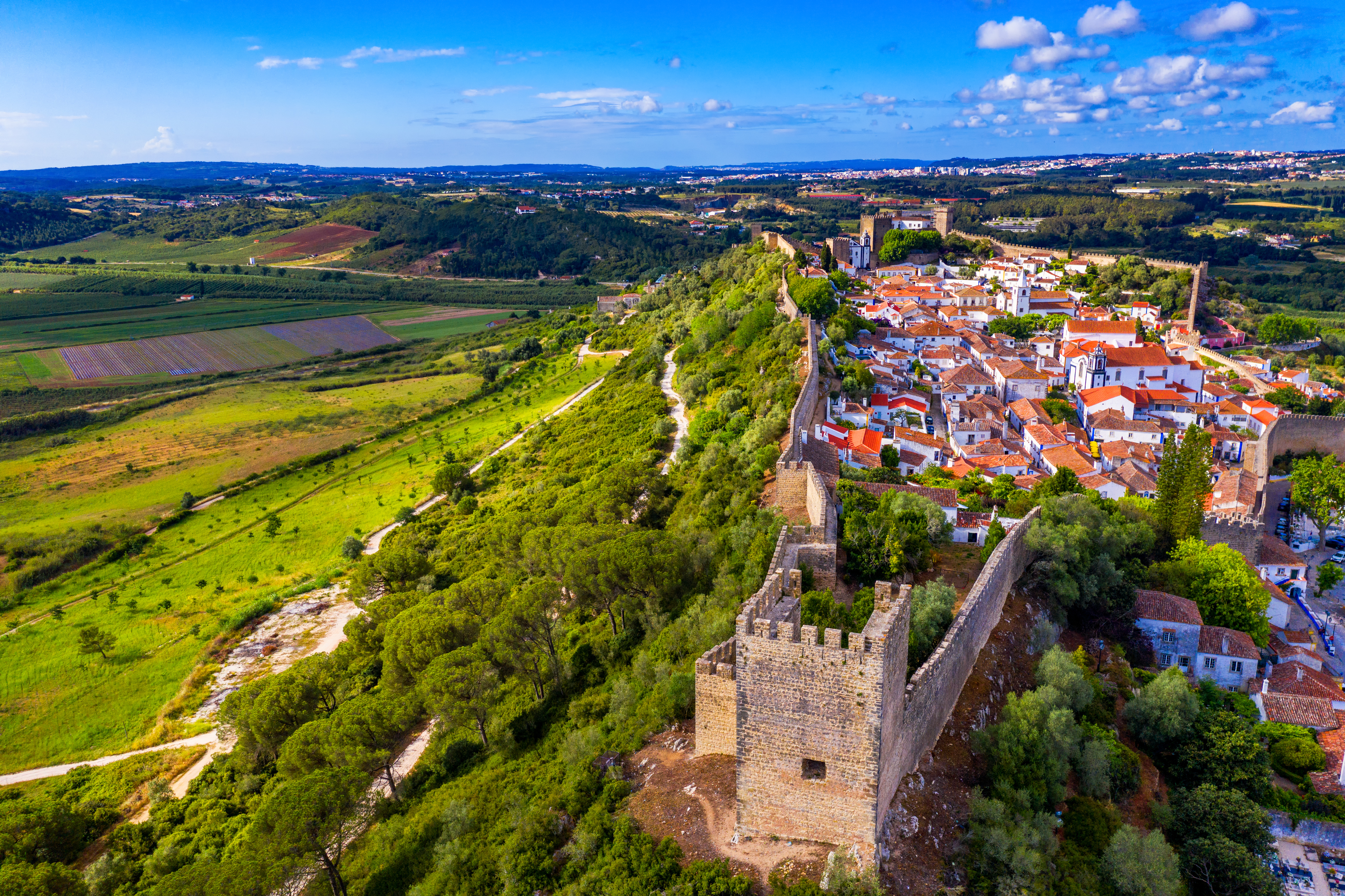 Luftfoto af den middelalderlige befæstede by Óbidos i Portugal med røde tegltage, gamle fæstningstårne og grønt landskab under en blå sommerhimmel