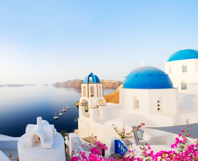 Traditionel græsk landsby Oia på Santorini med hvide huse og blå kupler mod det Ægæiske Hav og caldera omgivet af lyserøde blomster