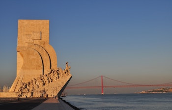Det imponerende Opdagelsesmonument i Lissabon badet i gyldent sollys, med den røde 25. April-bro over Tejo-floden i baggrunden