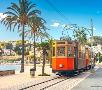 Ikonisk orange sporvogn på ruten mellem Soller og Port de Soller med palmer og blåt Middelhav i baggrunden på Mallorca