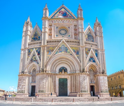 Imponerende panoramaudsigt af Orvieto-katedralen (Duomo di Santa Maria Assunta) i Umbrien, Italien, med dens storslåede gotiske facade prydet med farverige mosaikker og detaljerede skulpturer mod en klar blå himmel