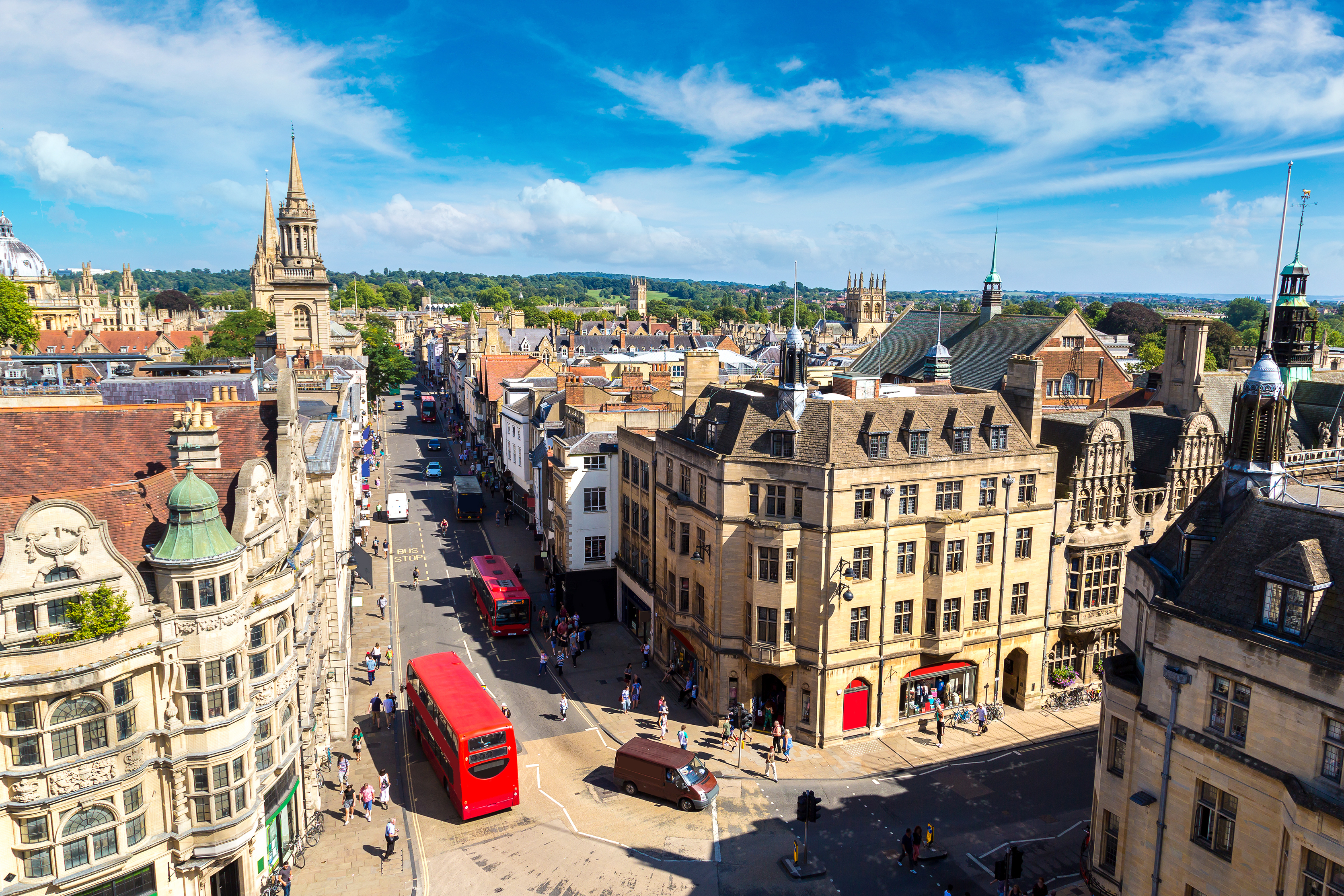 Luftfoto af Oxford med historiske universitetsbygninger, karakteristiske spir og en rød dobbeltdækkerbus på en solrig sommerdag i England