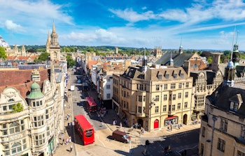 Luftfoto af Oxford med historiske universitetsbygninger, karakteristiske spir og en rød dobbeltdækkerbus på en solrig sommerdag i England