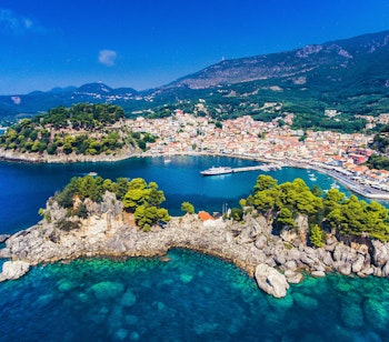 Luftfoto af den maleriske kystby Parga i Epirus-regionen med farverige bygninger mellem bjerge og det turkisblå Ioniske Hav med Panagia-øen og sandstrande
